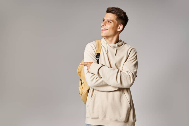 attraktiver junger Mann mit Rucksack und verschränkten Armen, der Seite mit Finger auf grauem Hintergrund zeigt - Foto, Bild