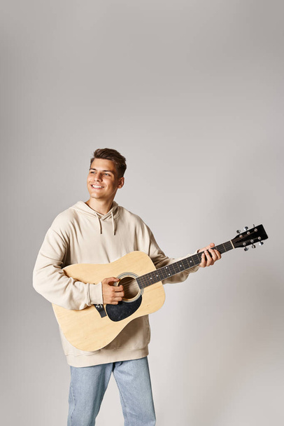 charming guy in his 20s with brown hair playing on guitar and smiling against light background - Photo, Image