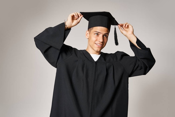 attractive student in gown with grey eyes holding graduate cap on head in light background - Photo, Image