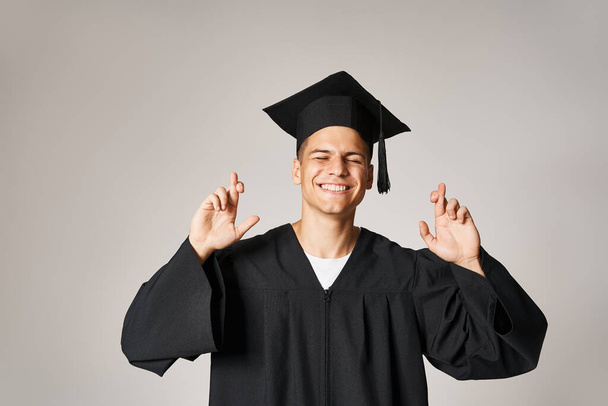 pohledný student v absolventských šatech a čepice přání se zavřenýma očima na šedém pozadí - Fotografie, Obrázek