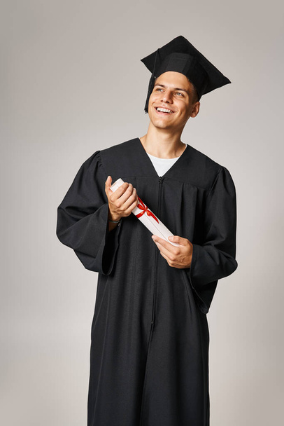 aantrekkelijke student in afgestudeerde jurk en cap vasthouden aan het diploma met de handen en op zoek naar toekomst - Foto, afbeelding