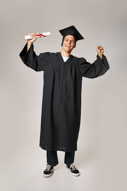 happy handsome student in graduate gown and cap rejoices in receiving diploma on grey background - Photo, Image