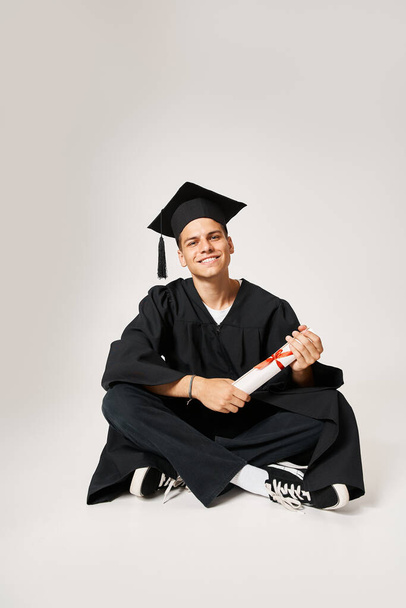 hombre guapo en vestido de graduado y gorra sentado y sosteniendo el diploma con las manos sobre fondo gris - Foto, Imagen