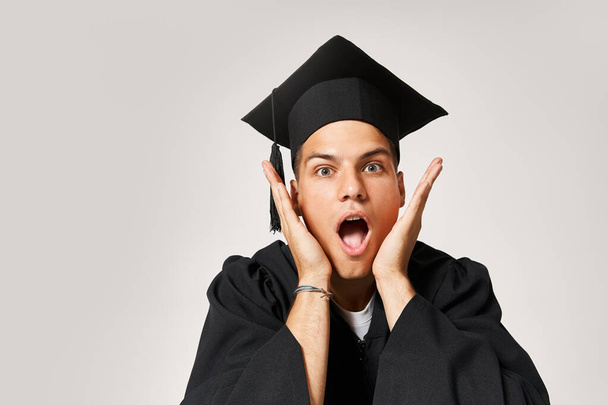 retrato de estudiante sorprendido en gorra de graduado mirando a la cámara sobre fondo gris - Foto, imagen