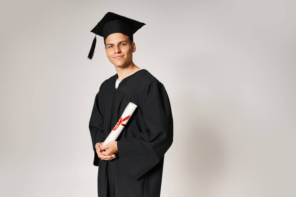 charismatic young student in graduate outfit holding to diploma with hands on grey background - Photo, Image