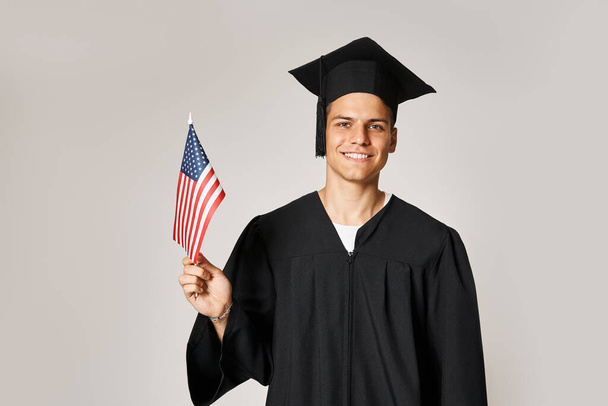 schöner junger Mann im Diplom-Outfit posiert mit amerikanischer Flagge mit Hand auf grauem Hintergrund - Foto, Bild