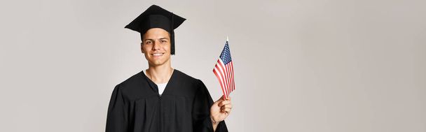 πανό του φοιτητή με στολή απόφοιτου ποζάρουν με αμερικανική σημαία με το χέρι σε γκρι φόντο - Φωτογραφία, εικόνα
