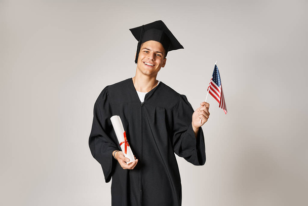 alegre hombre en traje de graduado posando con bandera americana y diploma con las manos sobre fondo gris - Foto, imagen