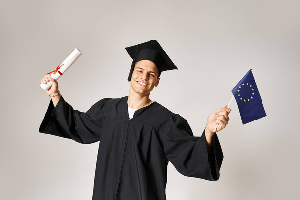 europäischer Student im Diplom-Outfit glücklich, sein Studium auf grauem Hintergrund abgeschlossen zu haben - Foto, Bild