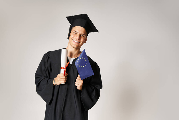attractive guy in graduate outfit posing with diploma and european flag in hands on grey background - Photo, Image