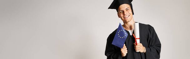 Banner eines Mannes im Diplom-Outfit posiert mit Diplom und Europafahne in den Händen auf grauem Hintergrund - Foto, Bild