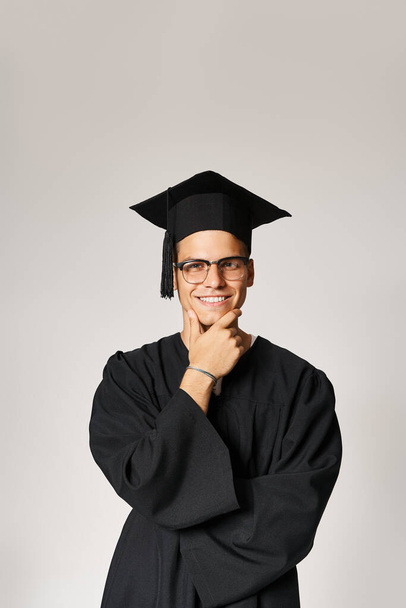 étudiant souriant en tenue d'études supérieures et lunettes de vision touchant la main à la mâchoire sur fond gris - Photo, image