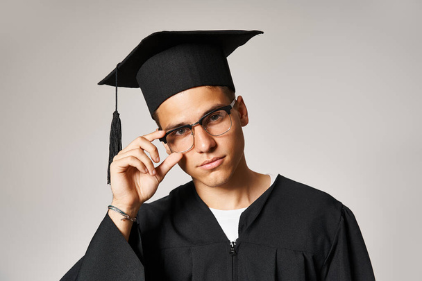 Красивый молодой человек в выпускном костюме трогает руки к очкам на сером фоне - Фото, изображение