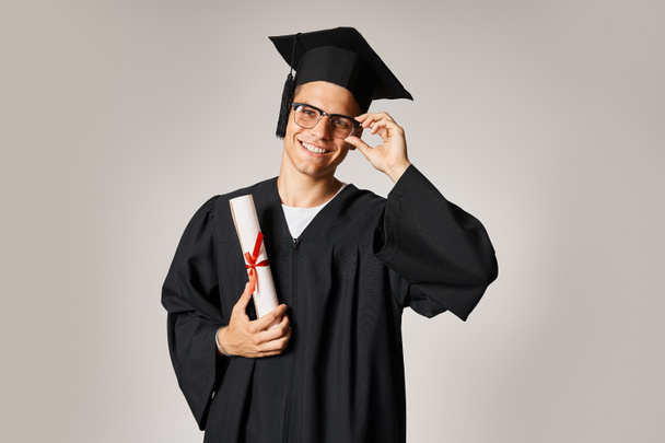 attractive student in graduate outfit touching hand to vision glasses and holding diploma - Photo, Image