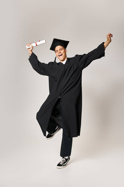vrolijke student in afgestudeerde outfit blij om zijn studies tegen grijze achtergrond hebben afgerond - Foto, afbeelding
