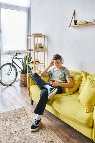 przystojny młody mężczyzna w słuchawkach siedzi na żółtej kanapie w domu i przewijanie do mediów społecznościowych - Zdjęcie, obraz