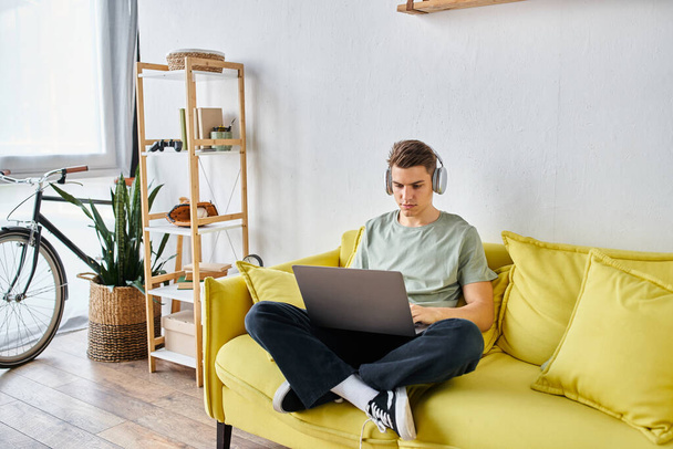 skupiony student z brązowymi włosami w słuchawkach w żółtej kanapie w domu ucząc się na laptopie - Zdjęcie, obraz