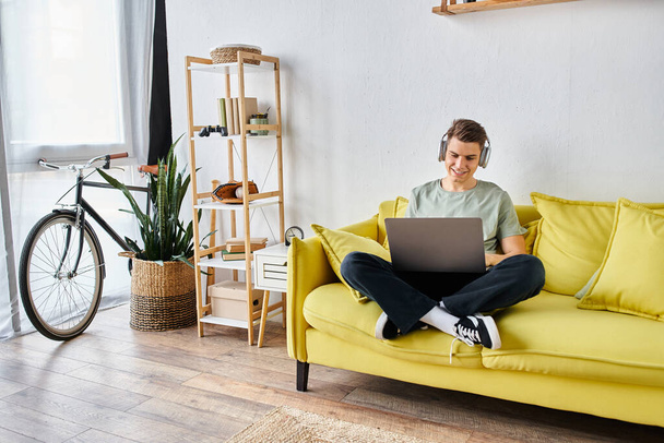 Улыбающийся привлекательный студент в наушниках в желтом диване дома учится на ноутбуке - Фото, изображение