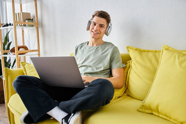 όμορφος νεαρός άνδρας με ακουστικά και φορητό υπολογιστή σε κίτρινο καναπέ στο σπίτι κοιτάζοντας προς την κάμερα - Φωτογραφία, εικόνα