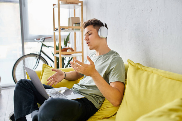 étudiant concentré avec écouteurs dans le canapé jaune à la maison parlant à une réunion en ligne dans un ordinateur portable - Photo, image