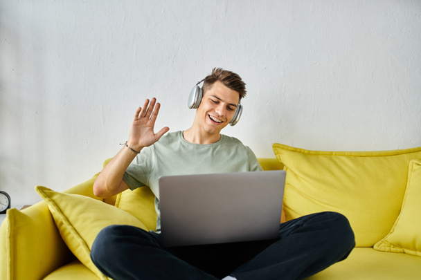 όμορφος νεαρός άνδρας με ακουστικά και laptop σε κίτρινο καναπέ λέγοντας γεια σε απευθείας σύνδεση συνάντηση - Φωτογραφία, εικόνα