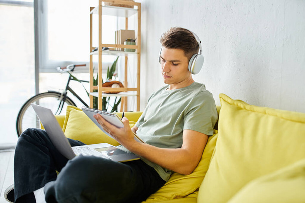 encantador joven estudiante con auriculares y portátil en sofá amarillo estudiar y escribir en nota - Foto, imagen