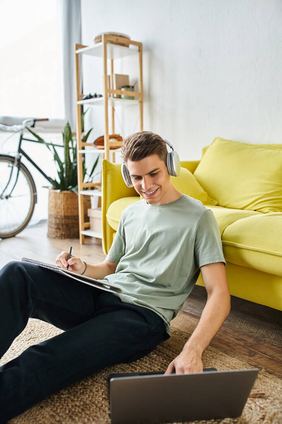 cheerful guy with headphones on floor near yellow couch studying in laptop and writing in note - Фото, зображення