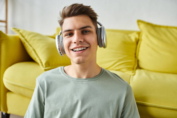 πορτρέτο του χαμογελαστού γοητευτικός νεαρός άνδρας στα 20 του με ακουστικά κάθεται κοντά σε κίτρινο καναπέ - Φωτογραφία, εικόνα