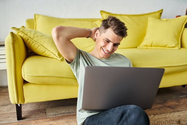 confundido estudiante en el suelo cerca de sofá amarillo en casa estudiando en el ordenador portátil y poniendo la mano detrás de la cabeza - Foto, imagen