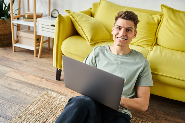 charming young man on floor near yellow couch at home sitting with laptop and smiling to camera - Photo, Image