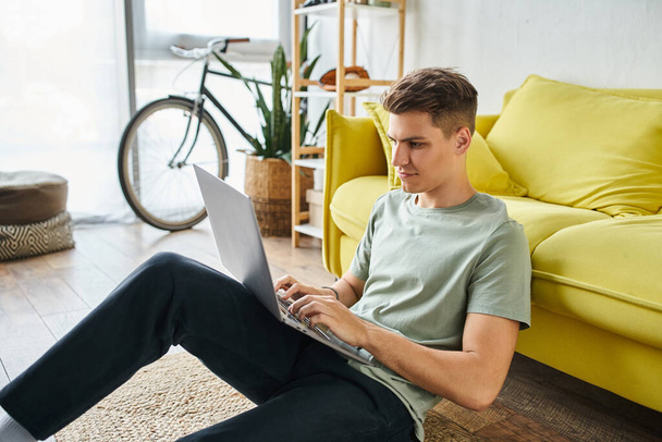 atractivo estudiante de 20 años en el suelo cerca de sofá amarillo en el hogar en red en el ordenador portátil de lado - Foto, imagen