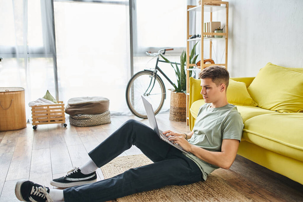 Student in den Zwanzigern mit braunen Haaren auf dem Boden neben gelbem Sofa zu Hause, der in Laptop seitlich studiert - Foto, Bild