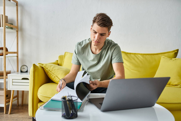 εστιασμένη μαθητής σε κίτρινο καναπέ στο σπίτι κάνει coursework με σημειώσεις και φορητό υπολογιστή στο τραπέζι του καφέ - Φωτογραφία, εικόνα