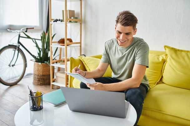 Улыбающийся парень в возрасте 20 лет на жёлтом диване дома делает курсовую работу с записками и ноутбуком на столе - Фото, изображение