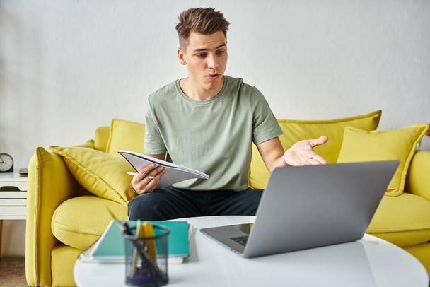 Verwirrter junger Student auf gelbem Sofa zu Hause bei der Kursarbeit in Notizen und Laptop auf Couchtisch - Foto, Bild