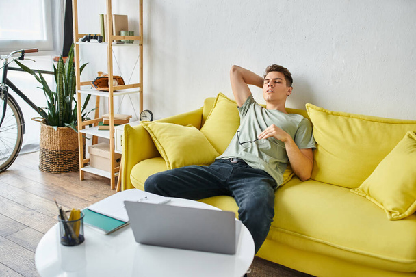Уставший парень с ноутбуком кладет руку за голову и опирается на желтый диван в очках - Фото, изображение