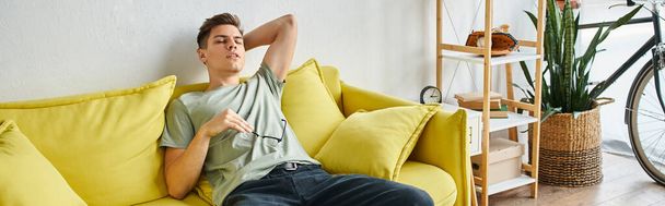 banner of tired young man putting hand behind head and leaning on yellow couch with glasses - Photo, Image
