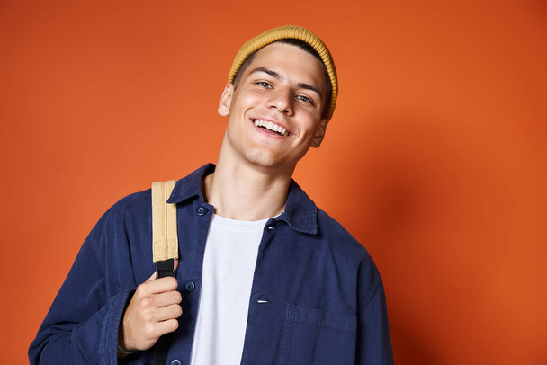 portrait of smiling handsome guy in yellow hat with backpack against terracotta background - Photo, Image
