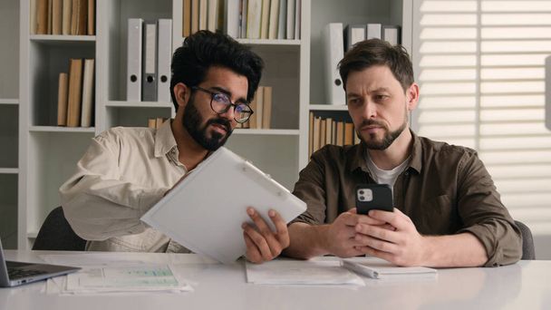 Két üzletember multiracionális férfi munkatársak irodai arab indiai üzletember show papírok üzleti projekt laptop kollégáról kollégára kaukázusi férfi figyelmen kívül hagyja függő mobiltelefon hatástalan csapatmunka - Fotó, kép