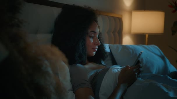 Hezká africká žena použít smartphone ležící v posteli noc nespavost relaxaci v domácí ložnici mobilní telefon rolování mediální sítě usmívající se žena blogger vlivnější dívka gadget narkoman před spaním - Záběry, video