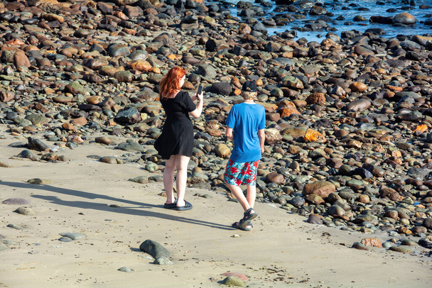 Молодая женщина в коротком черном платье стоит на каменистом пляже и фотографирует на мобильный телефон, рядом с ней стоит мальчик - Фото, изображение