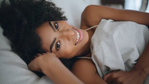Happy lady wakeup bedroom portrait. Healthy woman relaxing in comfortable bed closeup. African carefree girl looking camera lying at mattress alone. Smiling model on soft pillow. Morning awake concept - Photo, Image
