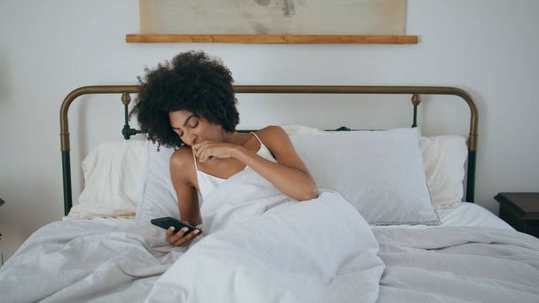 Heräävä tyttö haukottelee sängyssä aamulla. Rento nainen katsomassa puhelimen lukemista uutisia valkoisessa makuuhuoneessa. Unelias afrikkalainen nainen makaa tyyny kädessä kännykkä laite. Laiska malli surffailu puhelin app viikonloppuna - Valokuva, kuva