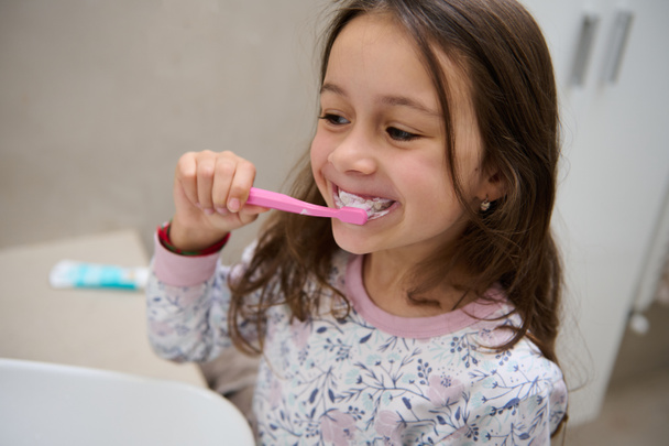 Adorabile bambina in pigiama, che tiene uno spazzolino da denti con dentifricio e si lava i denti bianchi davanti allo specchio nel bagno di casa. Uno stile di vita sano. Cura orale dentale e igiene - Foto, immagini