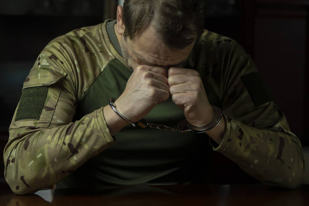 Солдат в камуфляже, руки в наручниках, за столом в помещении, темный тон. - Фото, изображение