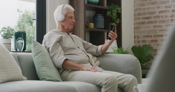 Écouteurs, maison et homme âgé avec un smartphone, musique en streaming et écoute de la radio dans un salon. Pensionné dans un salon, appartement et vieil homme avec un téléphone portable, casque et audio avec application. - Séquence, vidéo