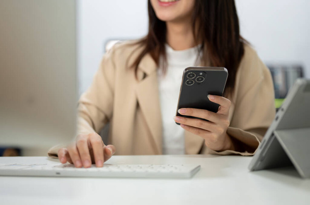 Przycięty obraz azjatyckiej biznesmenki używającej smartfona i wpisującej na klawiaturze komputer przy biurku w biurze. Koncepcje ludzi i technologii - Zdjęcie, obraz