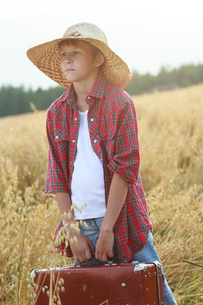 Teenage traveler in farm oat field holding old-fashioned suitcase and looking to horizon - Foto, Imagen