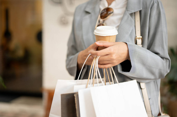 Gesneden beeld van een vrouw in trendy kleding met boodschappentassen en een koffiebeker, wandelen buiten, genieten van winkelen in de stad. - Foto, afbeelding