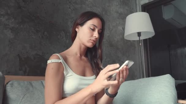 Woman sits on sofa in living room texting friends on phone Woman communicates ease embracing digital connections. Womans conversation flows through her device symbolizing modern social interaction - Footage, Video